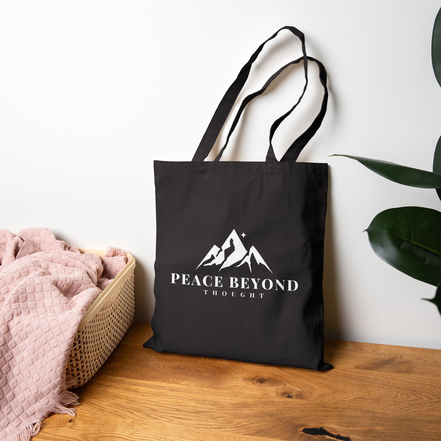 Black tote bag with mountain graphic and 'Peace Beyond Thought' text on wooden table