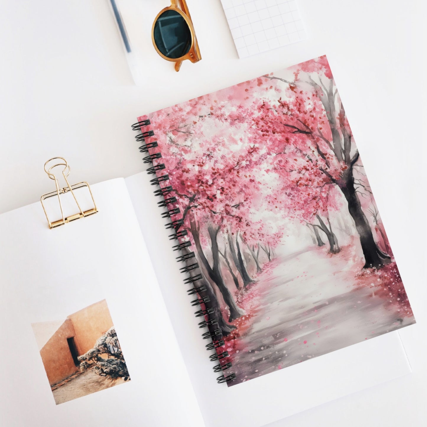 Spiral notebook with cover art of a peaceful pink blossoming trees alley on a white desk.
