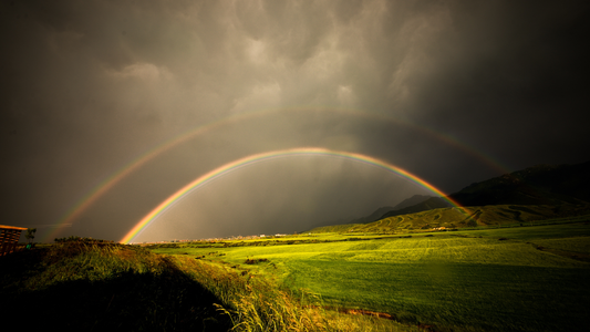 Double rainbow over green field and mountains under dramatic cloudy sky