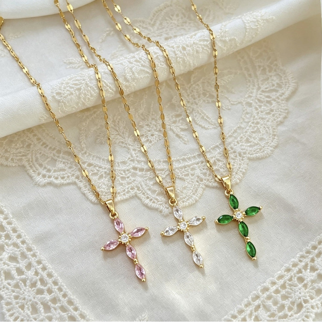 Pink, clear and green crystal cross pendants on separate gold-tone chains, displayed on pale lace