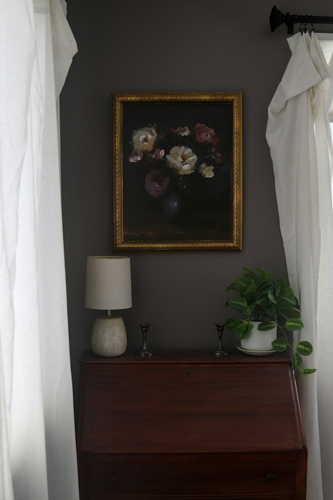 Dark floral painting above a wooden desk with lamp.