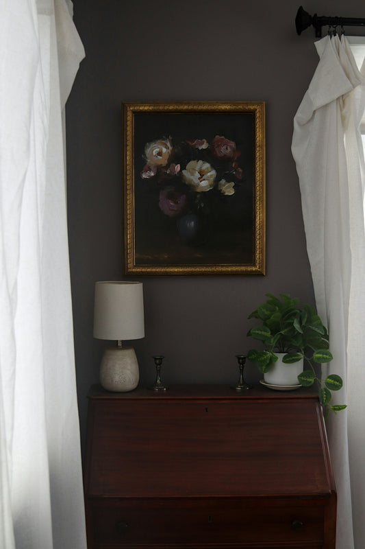 Dark floral painting above a wooden desk with lamp.