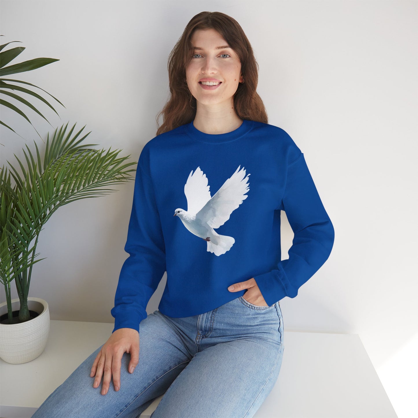 Woman in blue sweatshirt with white dove graphic, sitting by green potted plant