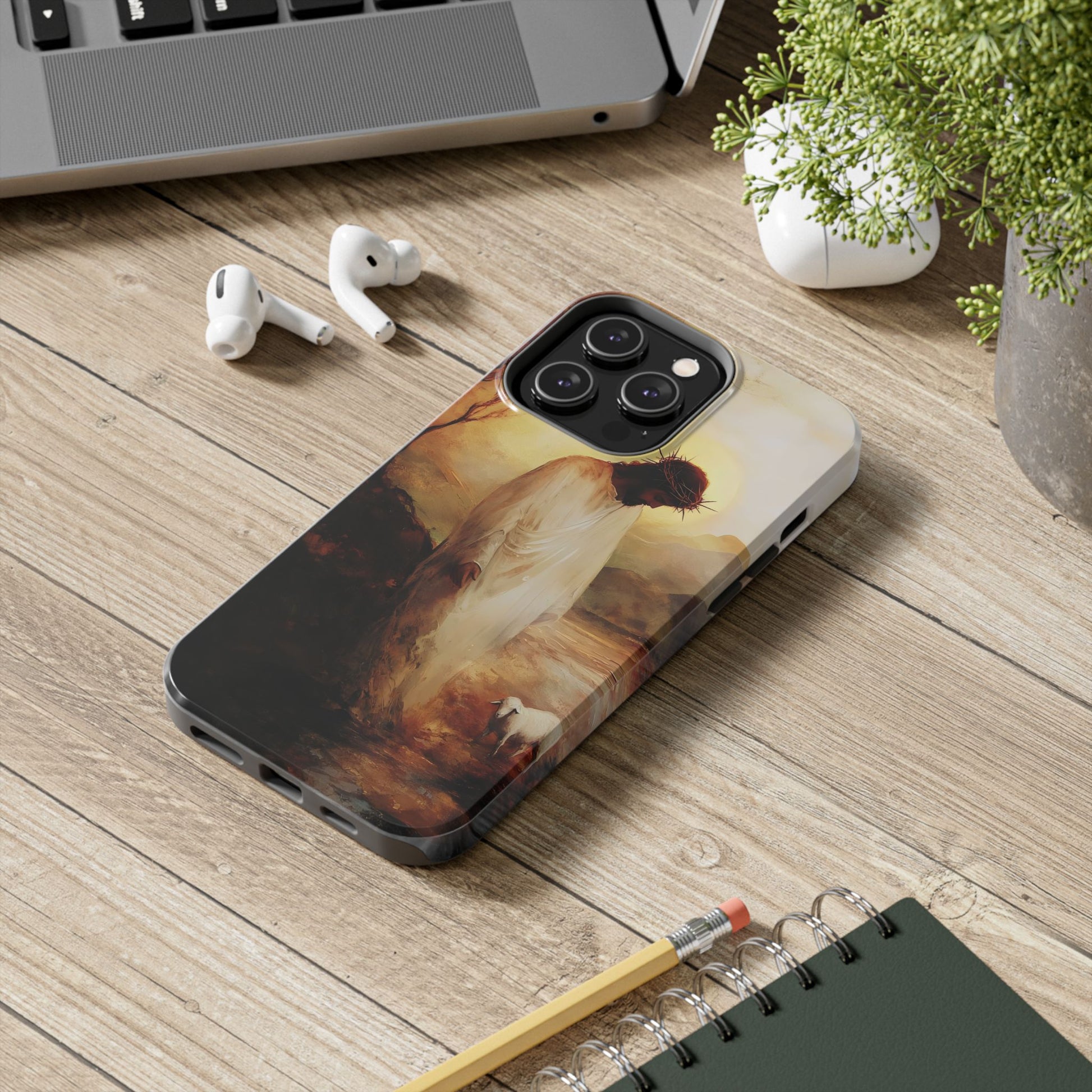 Christian phone case with Jesus and lamb artwork on wooden desk near laptop, earbuds, and plant