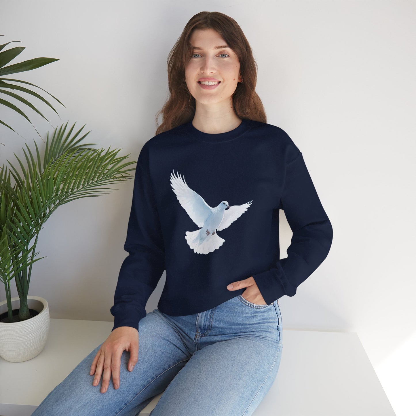 Woman wearing navy Christian sweatshirt with white dove graphic, sitting indoors by plant