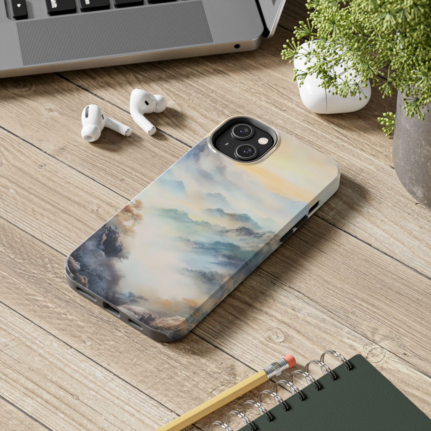 iPhone with misty mountain landscape case on wooden desk near laptop, earbuds, and plant