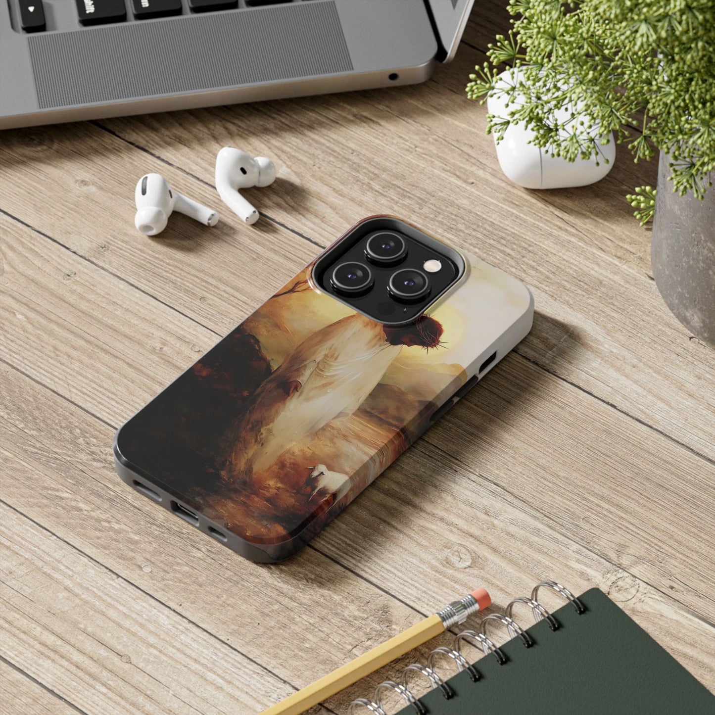 Christian phone case with Jesus and lamb design on wooden desk near laptop, earbuds, plant
