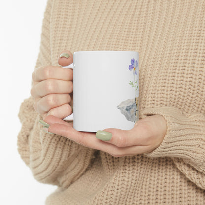 Woman in beige sweater holding white mug with purple flower and rock illustration