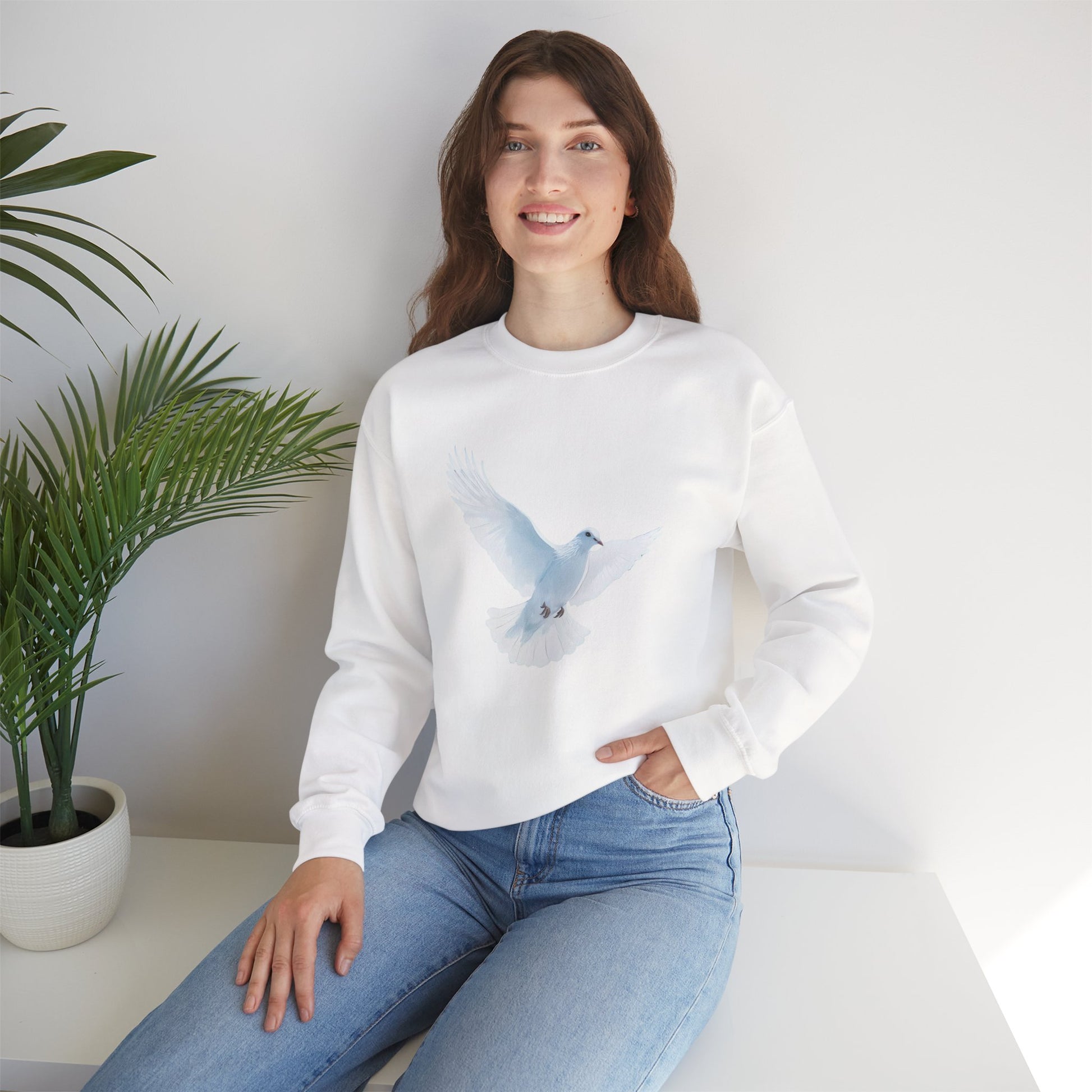 Woman in white sweatshirt with blue dove graphic, sitting indoors by a green plant