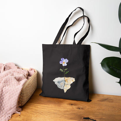 Black tote bag with a watercolor flower growing from cracked stone, on wooden surface