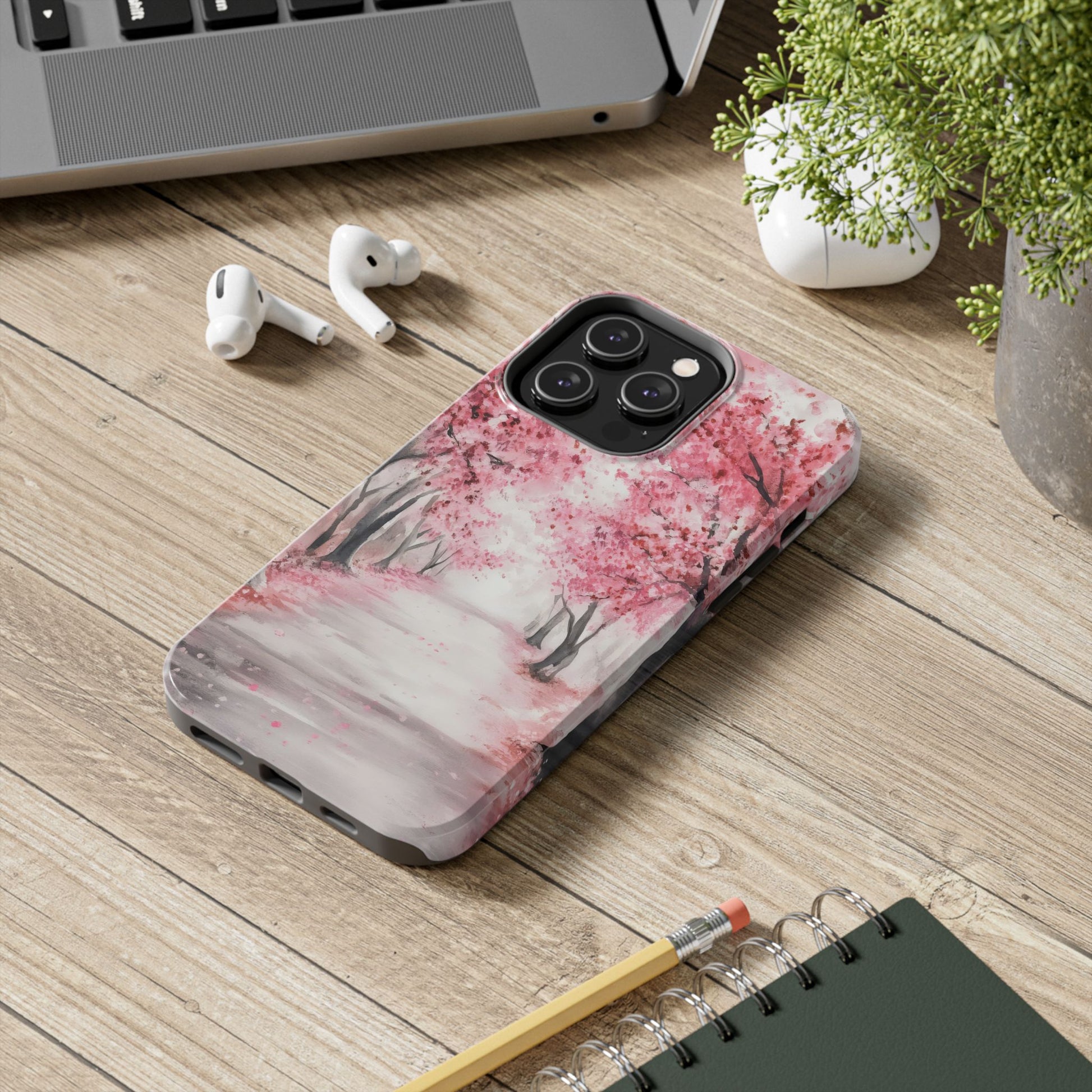 iPhone case with pink cherry blossom trees design on desk with AirPods and notebook