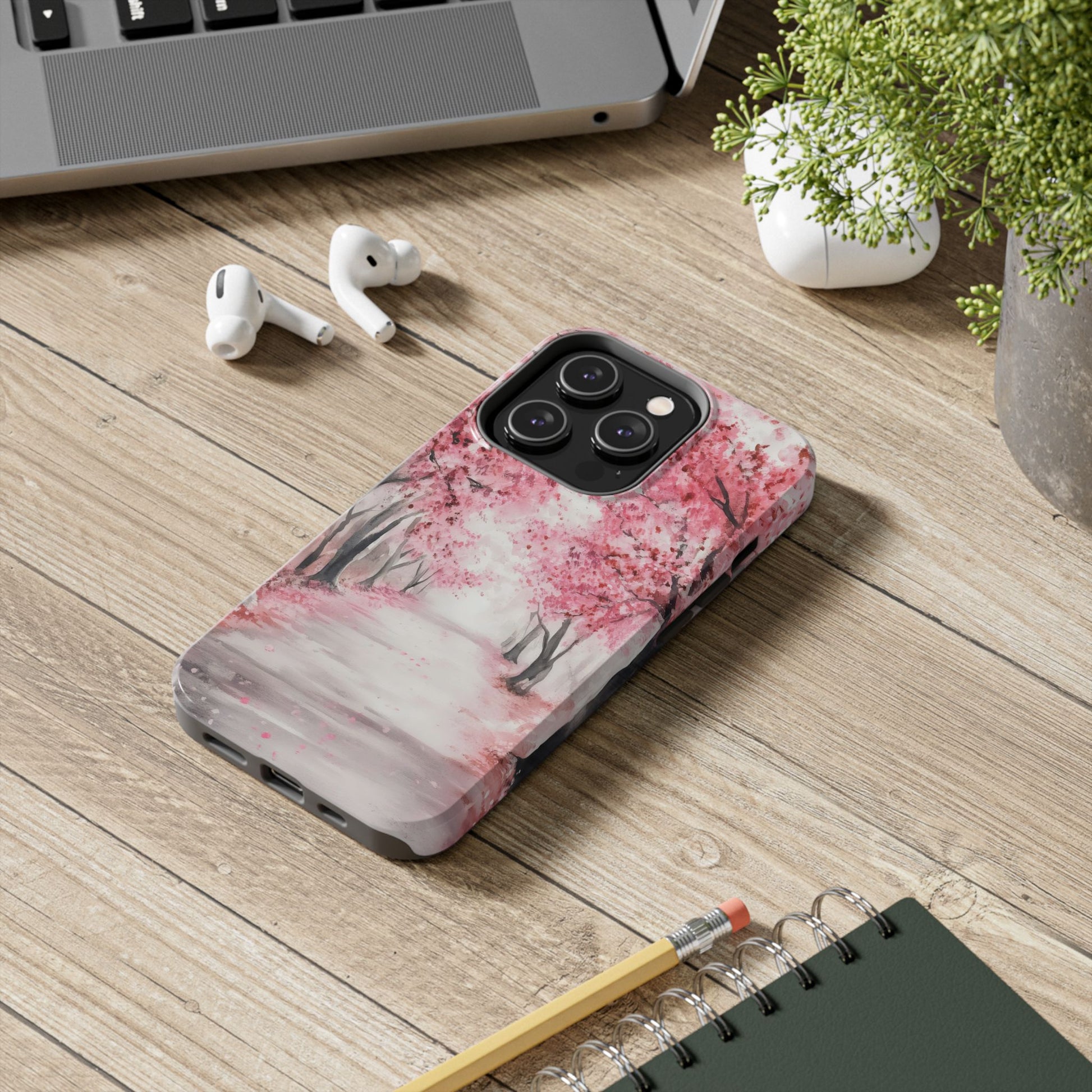 iPhone with pink cherry blossom tree case on wooden desk beside laptop, earbuds, plant, and notebook