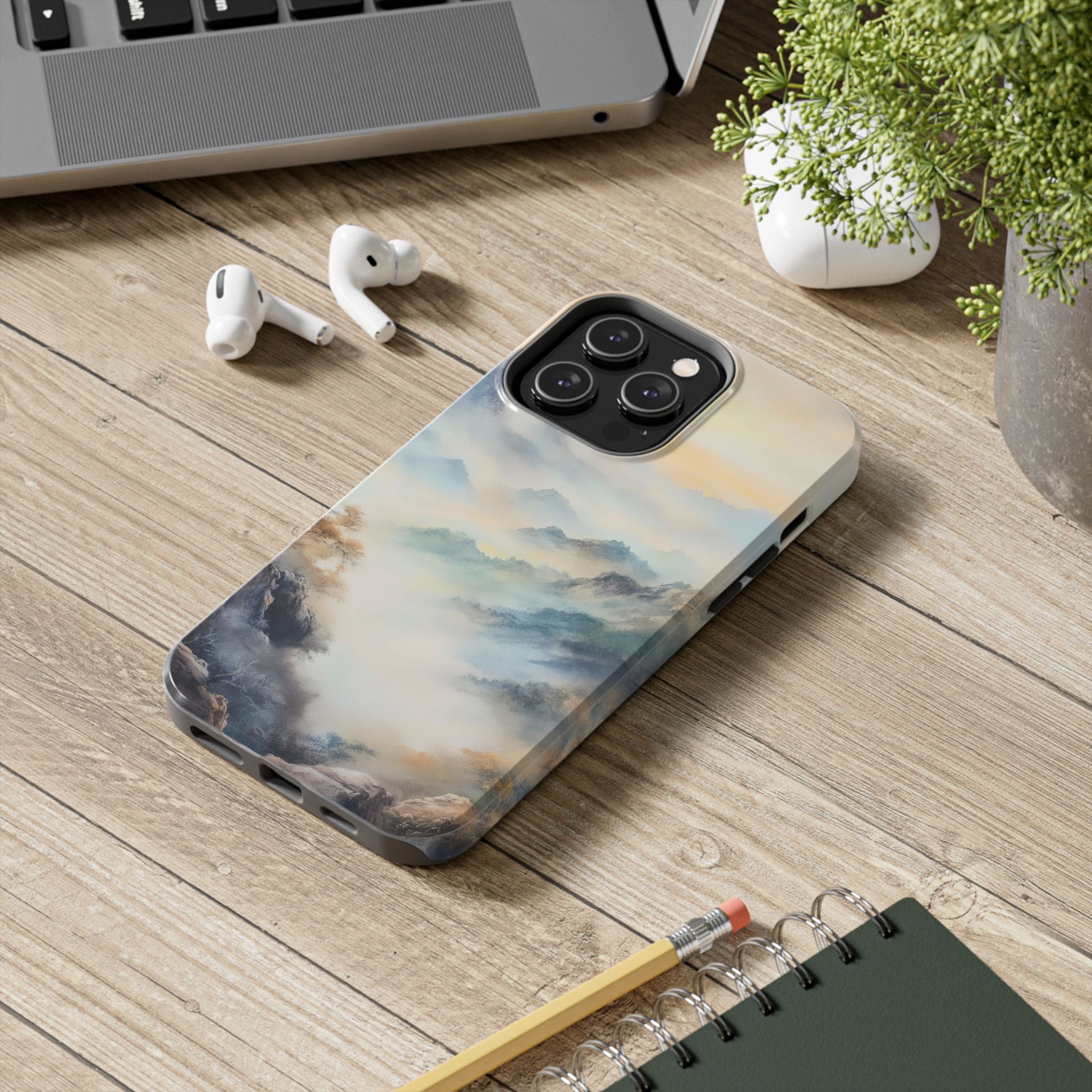 Phone case with serene mountain landscape art on wooden desk beside earbuds, notebook, and plant