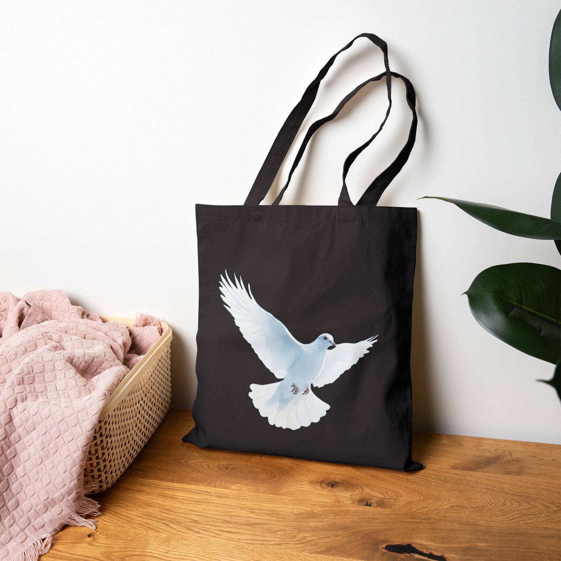 Black tote bag with white dove illustration, Christian gift, on wood table by blanket basket