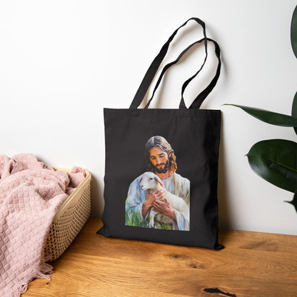 Black tote bag with artwork of Jesus holding a lamb, displayed on wooden table near pink blanket