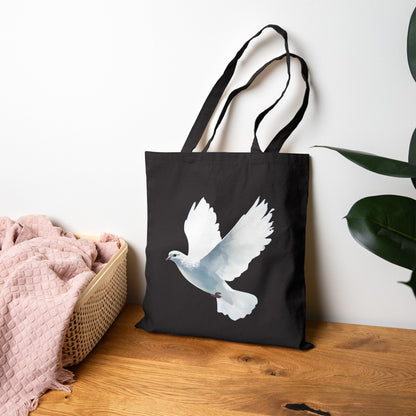 Black tote bag with white dove illustration, on wooden table near pink blanket and green plant