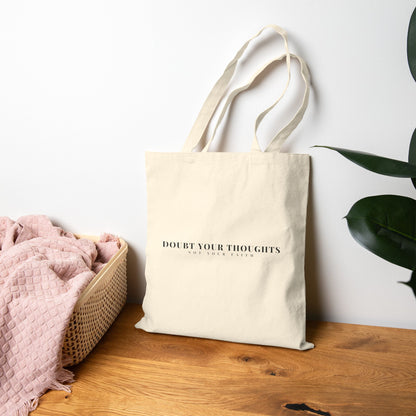 Beige tote bag with 'Doubt Your Thoughts Not Your Faith' text, on wood table near plant and basket.