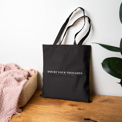 Black tote bag with 'Doubt Your Thoughts Not Your Faith' text, on wooden table near plant and pink blanket basket.