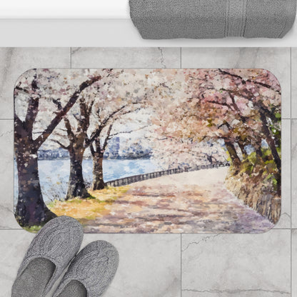 Bath mat with watercolor cherry blossom path by lake, gray slippers, and towel on tiled floor