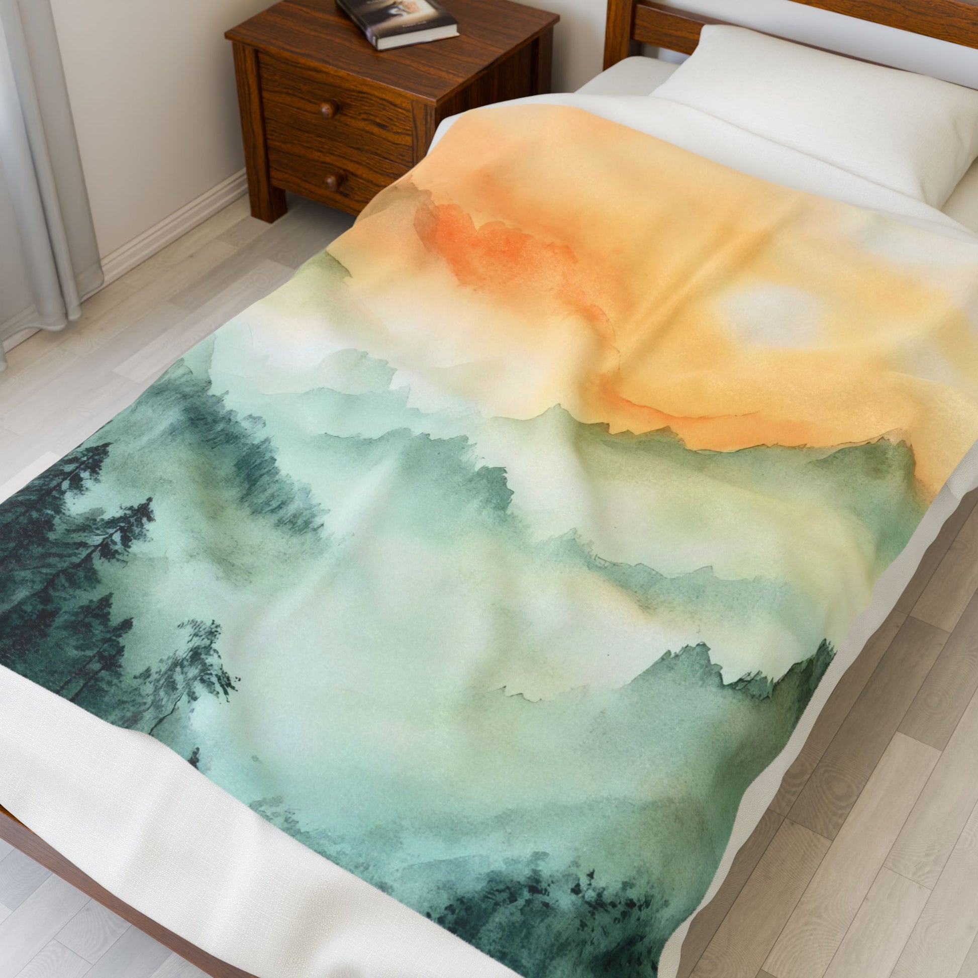 Soft blanket with watercolor mountain and sunrise design draped on a bed in a cozy bedroom.