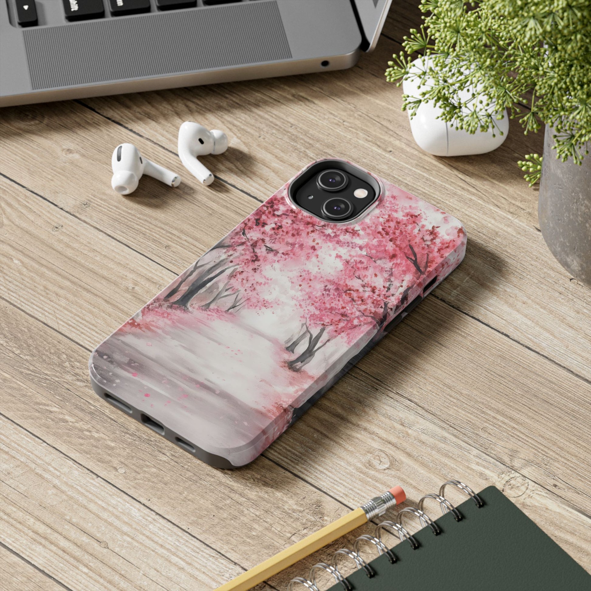 Phone case with pink cherry blossom tree art on wooden desk near notebook, pencil, and earbuds