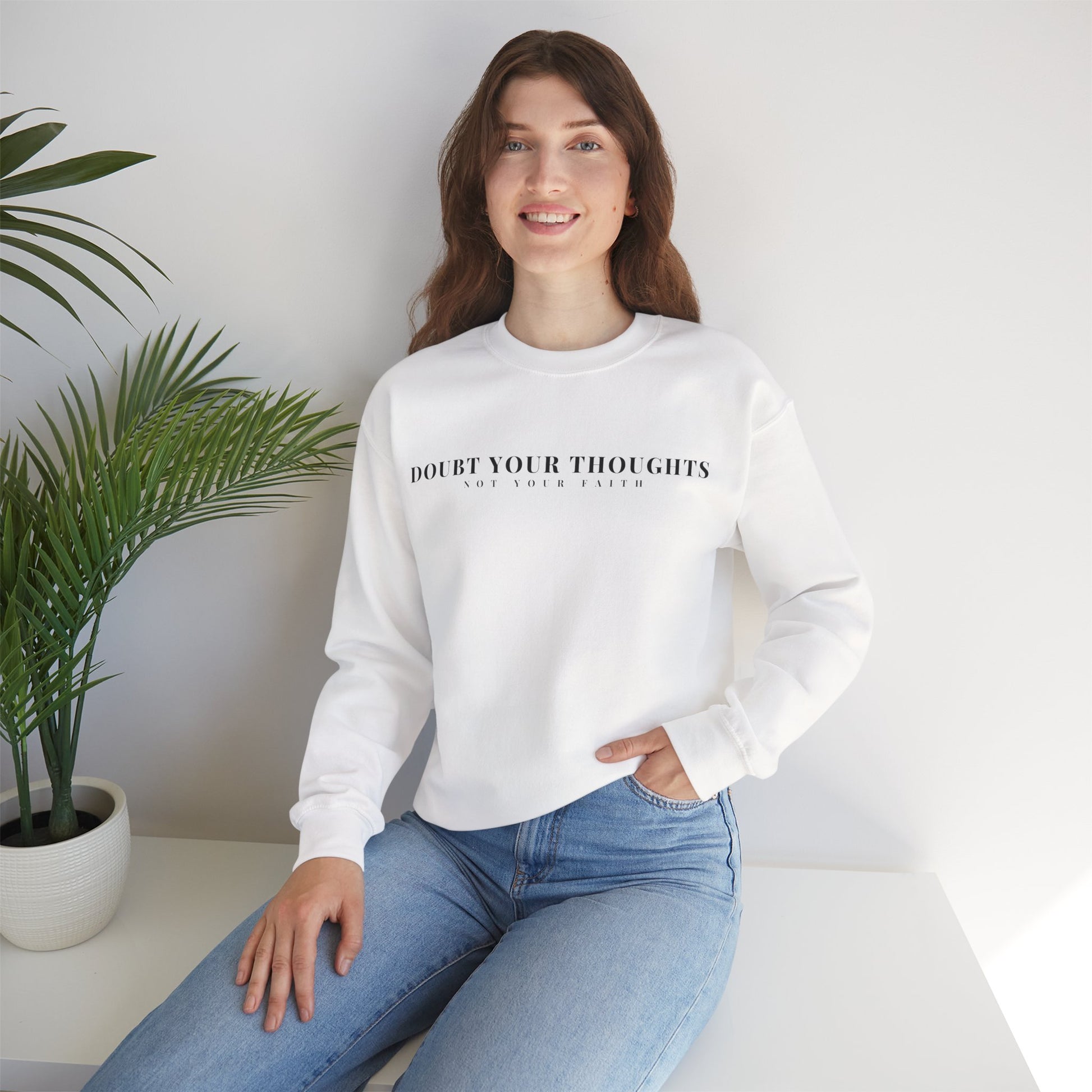 Woman in white sweatshirt with 'Doubt Your Thoughts Not Your Faith' text, sitting by green plant.