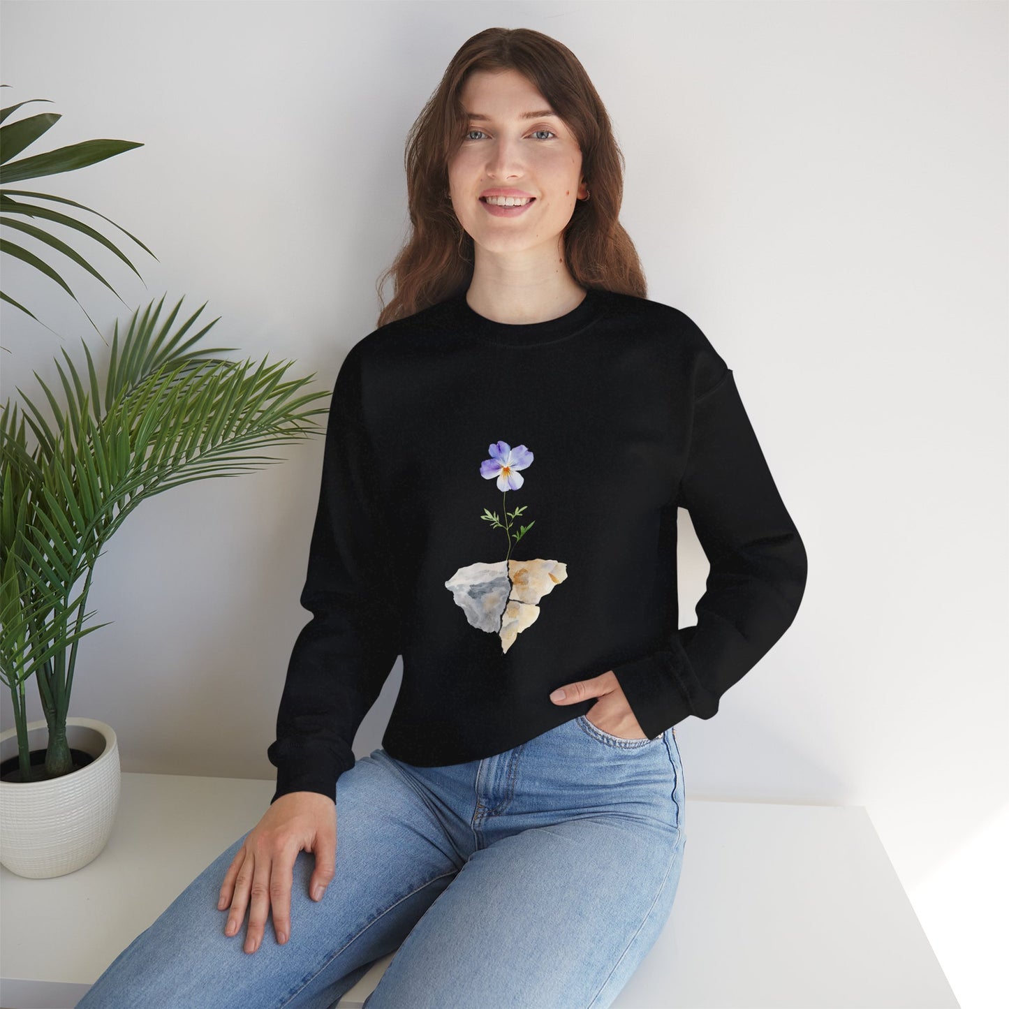 Woman wearing black sweatshirt with purple flower and rock graphic, sitting by potted plant