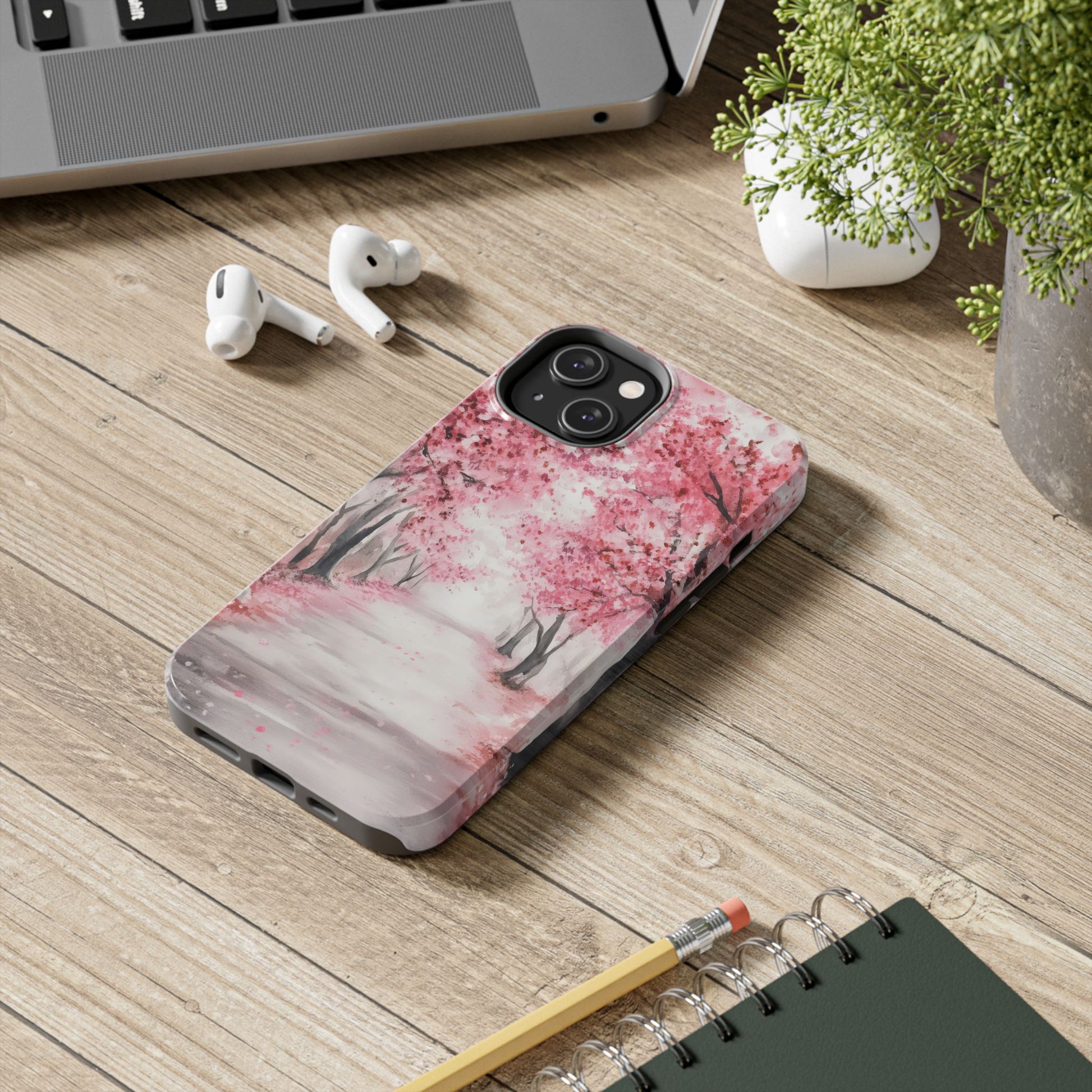 iPhone with pink cherry blossom tree art case on wooden desk near laptop and earbuds