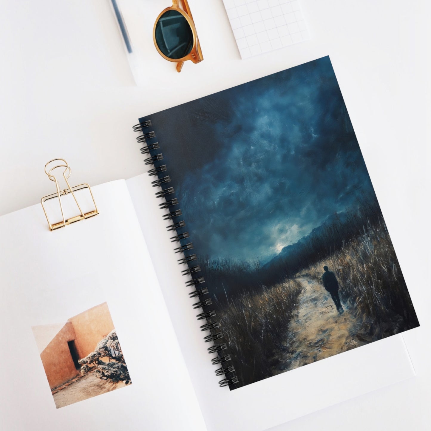 Spiral notebook with moody landscape painting of lone figure on path under cloudy sky, on minimalist desk