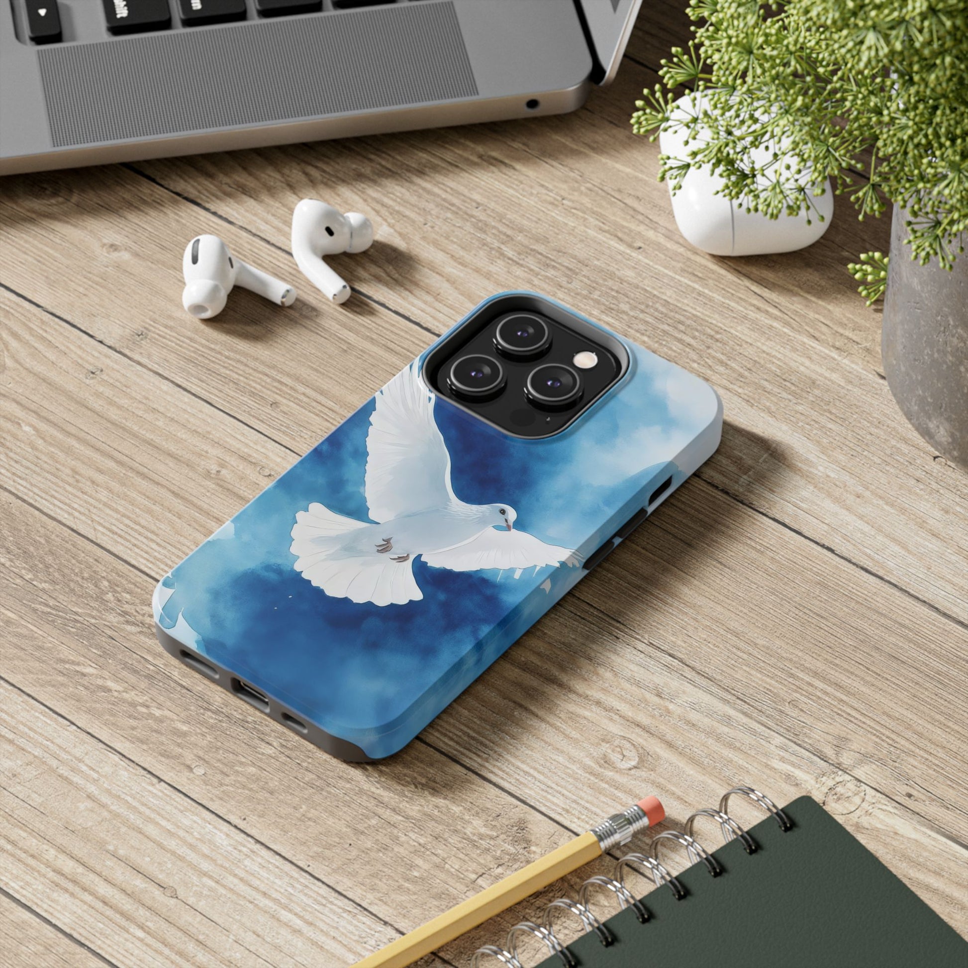 Phone case with white dove illustration on blue background, on wooden desk with laptop and earbuds